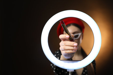 Beautiful woman holding lipstick through ring lamp on dark background, selective focus. Space for textの写真素材
