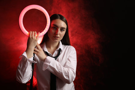 Beautiful woman with ring lamp on dark red background in smoke, space for text