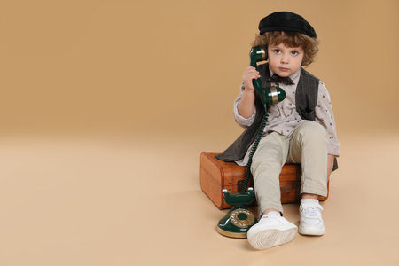 Cute little boy with old telephone and suitcase on beige background, space for text
