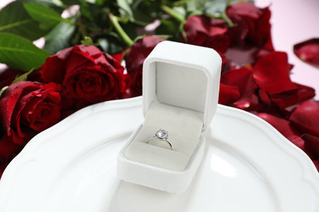 Beautiful setting for romantic dinner, engagement ring and rose petals on light table, closeup