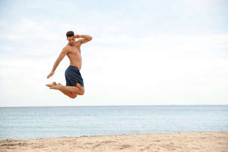 Muscular man jumping on beach, space for text. Body training
