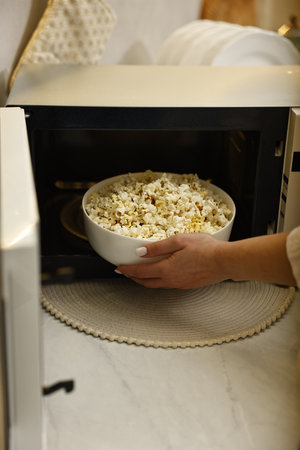 Woman taking bowl with popcorn out of microwave oven at white marble countertop in kitchen, closeupの写真素材