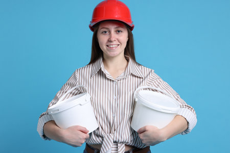 Woman wearing hardhat with buckets of paint on light blue backgroundの写真素材