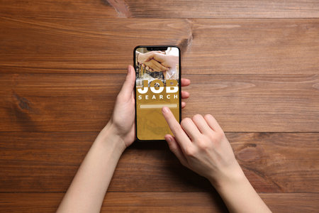 Job search. Woman using employment website on mobile phone at wooden table, top view