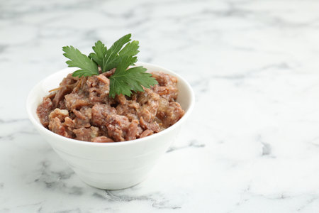 Canned meat with parsley in bowl on white marble table, closeup. Space for text