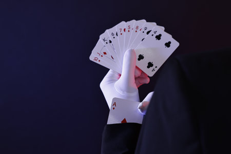 Illusionist hiding one playing card up his sleeve while showing deck on dark background, closeup....