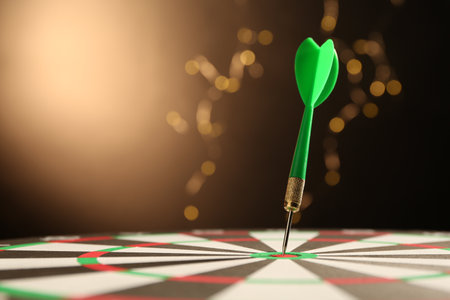 Green arrow hitting target on dart board against black background with blurred lights, closeup....