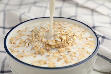 Pouring milk into bowl with oatmeal at table, closeup