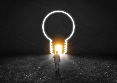Businessman walking towards light bulb neon outline and open door in dark stone wall. Idea, successful strategyの写真素材