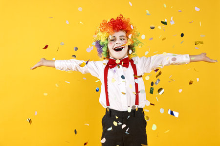 Happy little boy dressed like clown and flying confetti on yellow background. Surprise partyの写真素材
