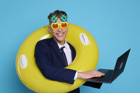 Businessman with inflatable ring, laptop and funny sunglasses on light blue backgroundの写真素材
