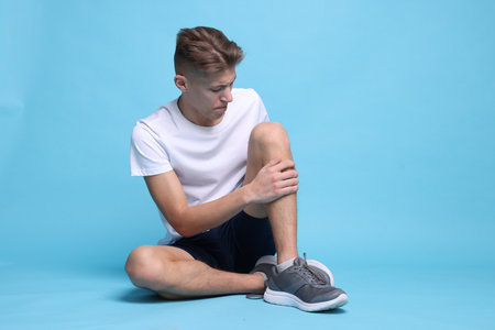 Young man suffering from pain in knee on light blue backgroundの写真素材