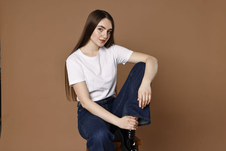 Beautiful young woman in stylish jeans sitting on stool against brown background