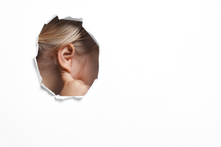 Little girl showing hand to ear gesture through hole in white paper, closeup.の写真素材