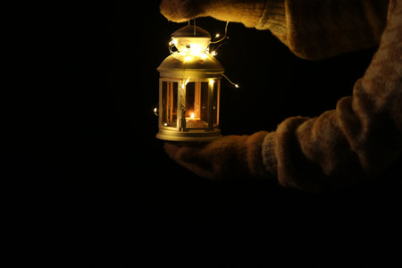 Woman holding Christmas lantern with burning candle and fairy lights in darkness, closeup. Space for...