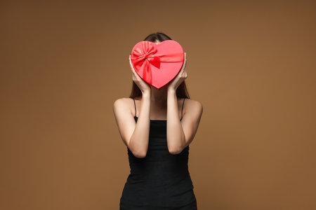 Happy Valentine's Day. Woman hiding behind heart shaped gift box on brown backgroundの写真素材
