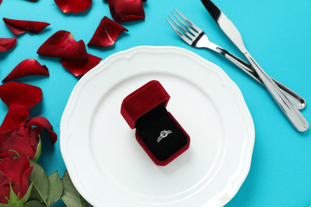 Beautiful setting for romantic dinner, engagement ring and rose petals on light blue background,...
