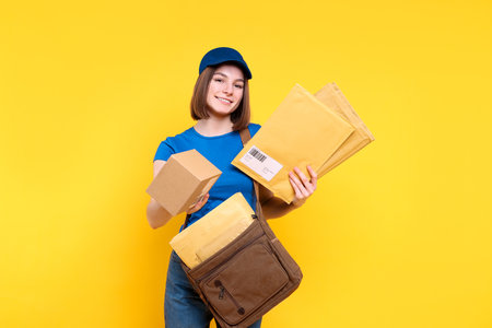 Happy postwoman with bag and envelopes giving parcel on yellow backgroundの写真素材