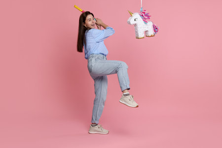 Happy woman hitting unicorn shaped pinata with stick on pink background
