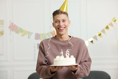 Coming of age party - 18th birthday. Happy young man in hat holding tasty cake with number shaped...