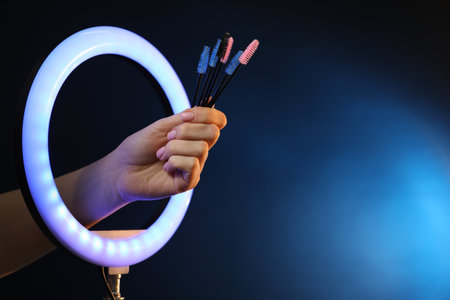 Woman holding makeup brushes through ring lamp on dark blue background, closeup. Space for textの写真素材