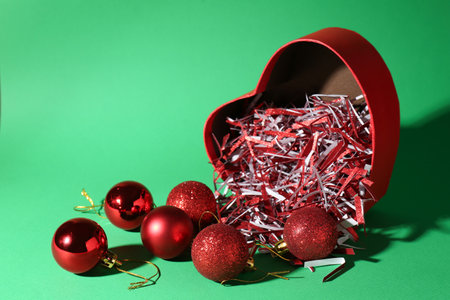 Red heart shaped box with Christmas balls and shiny filler on green background. Space for textの写真素材