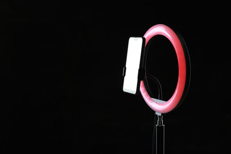 Ring lamp with smartphone on black background, space for text