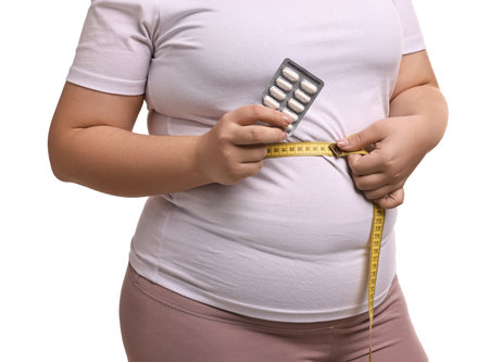 Plus size woman measuring waist with tape and holding blister of weight loss supplements on white background, closeupの写真素材