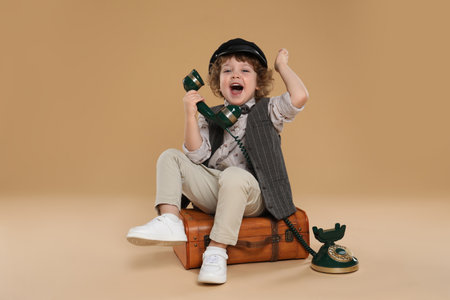 Cute little boy with old telephone and suitcase on beige backgroundの写真素材
