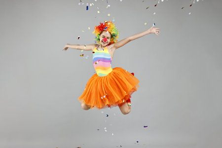 Girl dressed like clown jumping under flying confetti on grey background. Surprise partyの写真素材