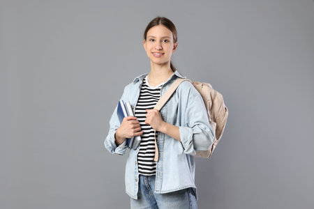 Portrait of smiling teenage girl with books and backpack on grey background