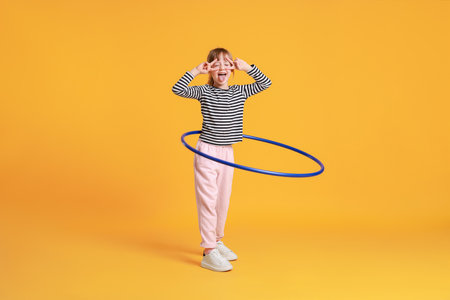 Cute little girl showing V-signs while exercising with hula hoop on orange background
