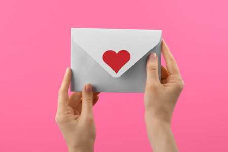 Woman holding envelope with heart sticker on pink background, closeup. Love letterの写真素材