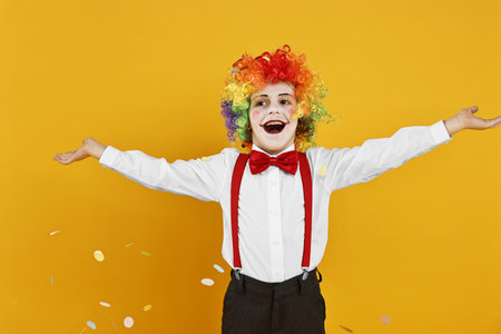 Happy little boy dressed like clown and flying confetti on yellow background. Surprise partyの写真素材