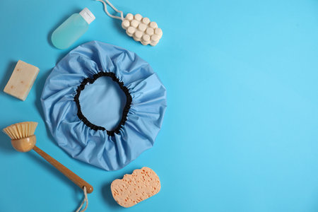 Shower cap and bath accessories on light blue background, flat lay. Space for text