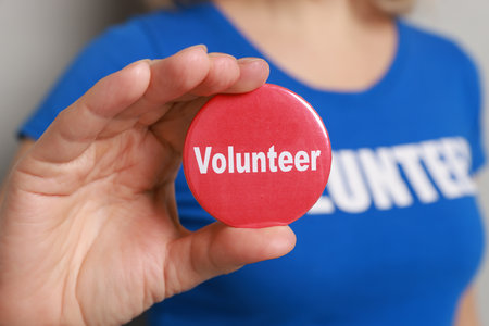 Woman holding button badge with word Volunteer on gray background, closeup