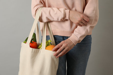 Woman holding shopper bag with groceries on light gray background, closeup. Eco friendly lifestyle