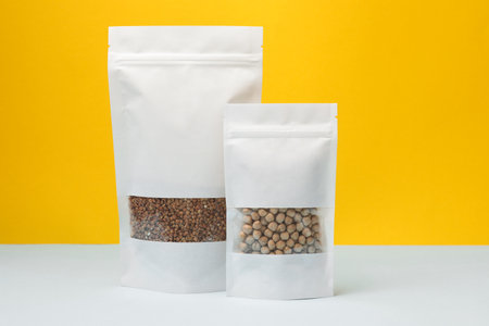 Paper pouch bags with chickpeas and buckwheat on table against yellow background