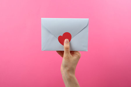 Woman holding envelope with heart sticker on pink background, closeup. Love letterの写真素材