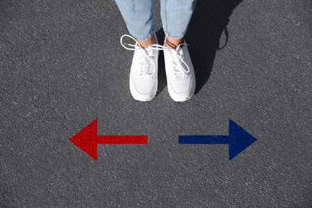 Woman standing near red and blue arrows pointing in different directions on asphalt road, closeup....
