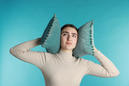 Distressed woman covering her ears with pillows from loud noise on blue backgroundの写真素材