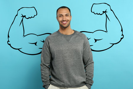 Handsome young man with drawing of strong arms behind him on light blue background