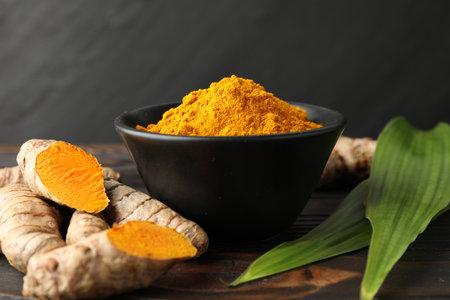 Aromatic turmeric powder in bowl, fresh roots and leaves on wooden table, closeup