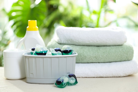 Container with laundry capsules, detergents and clean towels on table against blurred background,...