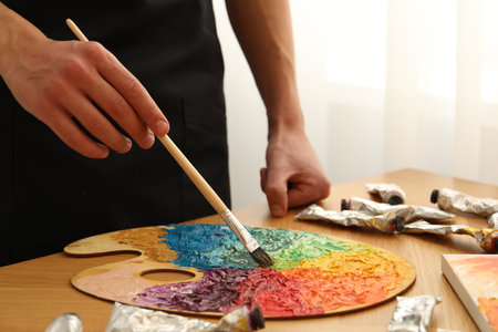 Man with paintbrush mixing paints on palette at wooden table indoors, closeupの写真素材