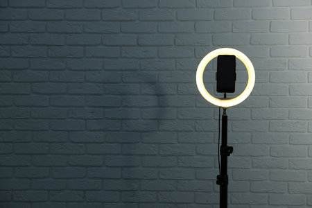 Ring lamp with smartphone against brick wall, space for text