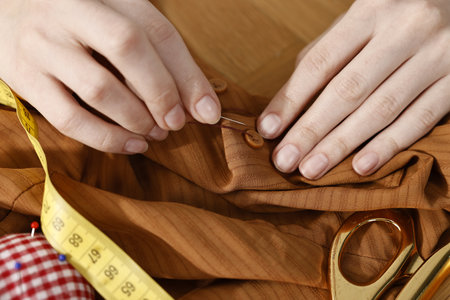 Woman sewing button with needle and thread onto brown shirt at table, above view
