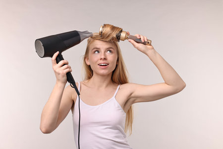Beautiful young woman styling her hair with hairdryer and brush on light grey backgroundの写真素材