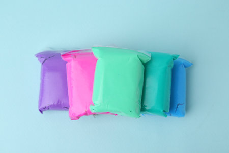 Different colorful play dough in packages on light blue background, top view