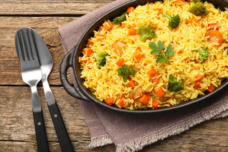 Tasty rice with vegetables in baking dish and cutlery on wooden table, top viewの写真素材